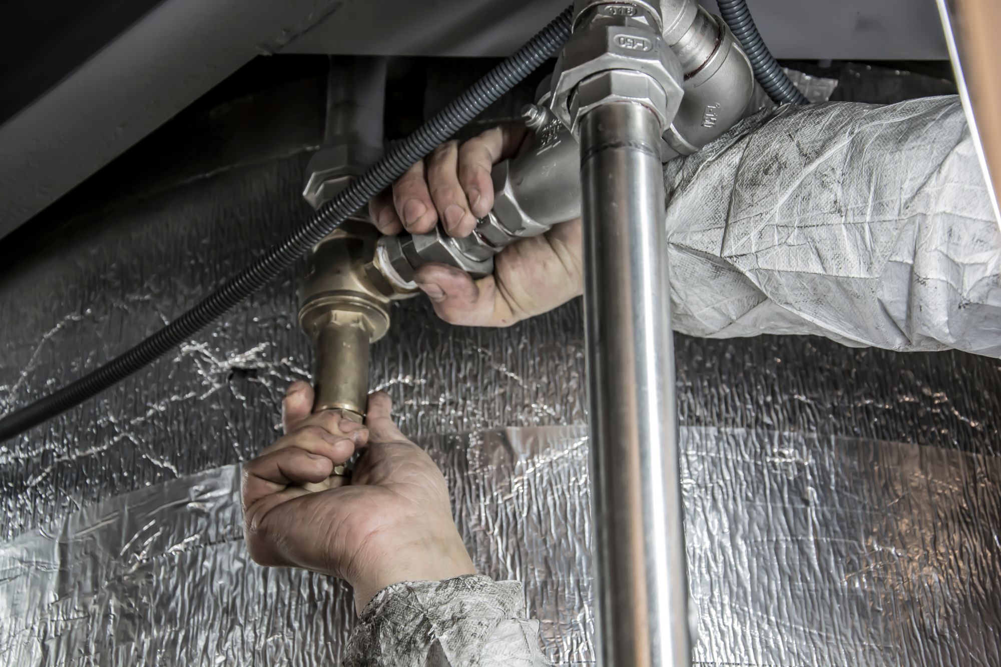A plumber's gloved hands tightening a steel pipe fitting under a floor